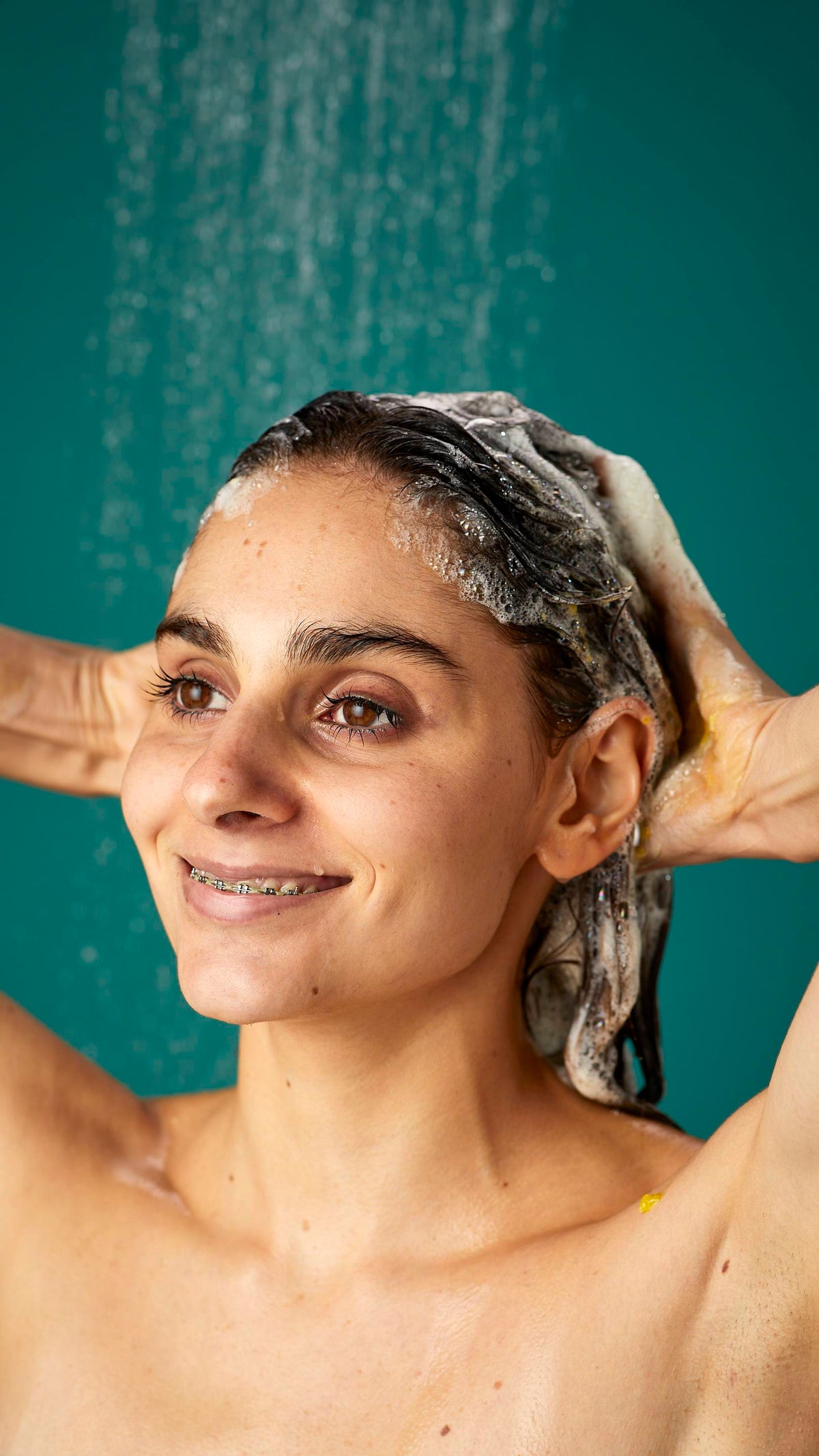 The model is standing under running shower water on a teal background. Their dark, long hair is wet and lathered in suds as they lather the Ginger shampoo into their roots.